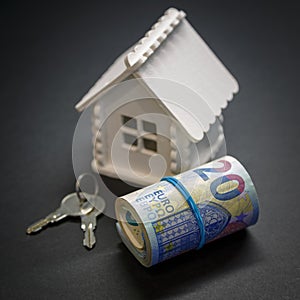 A roll consisting of euros, a house mock-up of white colour and keys to a future home on a black background