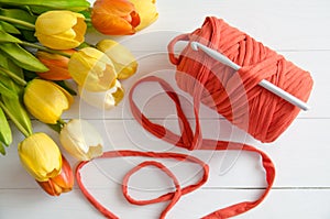 Roll of cotton yarn and a crochet hook with a bouquet of orange and yellow tulips on white wooden background.