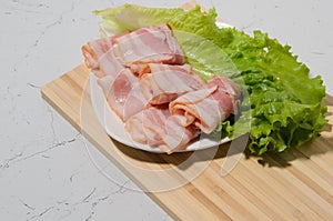 Rolled bacon slices are placed on a lettuce leaf on a plate. Bamboo cutting Board and texture table as background