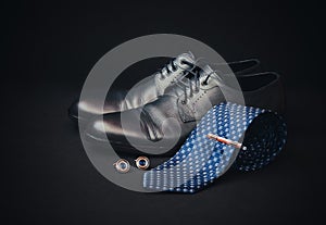 Rolled necktie, tie pin, cufflinks and black men`s shoes on dark background. Selective focus