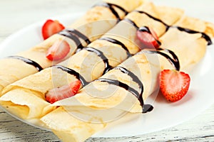 Rolled pancakes with strawberry on plate on white wooden background.