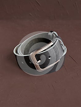 Rolled-up black fashionable men`s leather belt with metal buckle on brown background. Male accessory. Front view, vertical photo
