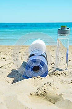 Rolled Yoga Mat Bottle with Water White Towel on Beach Sand with Turquoise Sea Blue Sky in Background. Sunlight