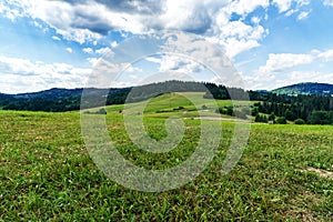 Zvlnená krajina s lúkami, zalesnenými kopcami a modrou oblohou s mrakmi