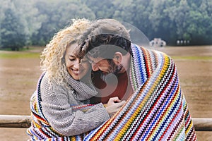 Romantic adult couple in love hug with tenderness under a colorful wool cover in outdoor park. Concept of winter leisure activity