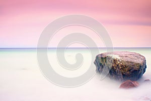 Romantic atmosphere in peaceful morning at sea. Big boulders sticking out from smooth wavy sea. Pink horizon