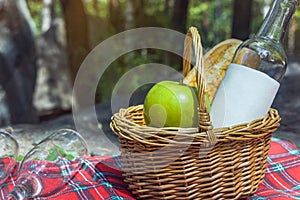 Romantic autumn picnic lunch outdoors. Wicker basket with baguette,   bottle of rose wine and apples on checkered cloth on stones