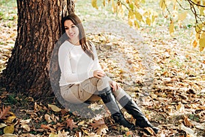 Romantic autumnal portrait.Beautiful brunette stylish girl is sitting near a big tree in the autumn park.Bright sunny day.Good