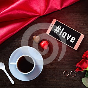 Romantic background with wedding ring, rose flowers, smartphone, cup of coffee and chocolate candy. Hashtag love