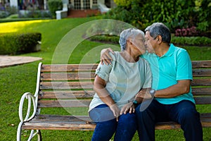 Romantic biracial senior husband kissing senior woman while sitting on bench in park