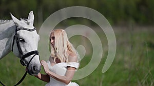 romantic blonde in white wedding dress is communicating with horse at nature, portrait shot