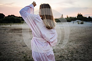 Romantic blonde woman in pink robe on the beach by the sea at sunset.