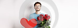 Romantic boyfriend waiting for kiss, holding bouquet of roses flowers and big red heart on Valentines day, love in air