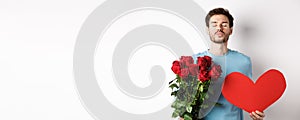 Romantic boyfriend waiting for kiss, holding bouquet of roses flowers and big red heart on Valentines day, love in air