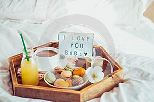 Romantic Breakfast in bed with I love you baby text on lighted box. Cup of coffee, juice, macaroons, flower and gift box on wooden