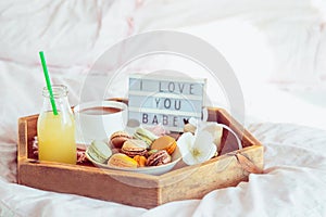 Romantic Breakfast in bed with I love you baby text on lighted box. Cup of coffee, juice, macaroons, flower and gift box on wooden