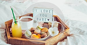 Romantic Breakfast in bed with I love you baby text on lighted box. Cup of coffee, juice, macaroons, flower and gift box on wooden