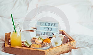 Romantic Breakfast in bed with I love you baby text on lighted box. Cup of coffee, juice, macaroons, flower and gift box on wooden