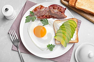Romantic breakfast with fried bacon, heart shaped egg and avocado toast on light grey table, flat lay. Valentine`s day celebratio