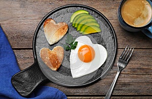 Romantic breakfast with fried heart shaped egg, avocado and toasts on wooden table, flat lay. Valentine`s day celebration