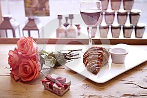 Romantic breakfast for Valentine`s Day celebrate. Present box, rose flowers, fresh croissant, wine on wooden table. Focus on crois
