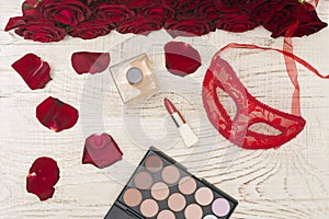 Romantic carnival concept. Red carnival mask, bouquet of red roses, lipstick, bottle of perfume and eye shadow on light wooden