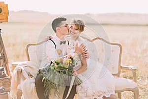 The romantic close-up portrait of the groom kissing the bride with the bouquet into the cheek in the sunny field. The