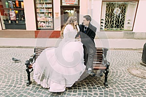 Romantic couple of bride and groom sitting on wooden bench in old town street
