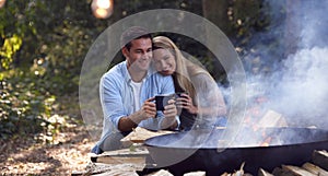 Romantic Couple Camping Sitting By Bonfire In Fire Bowl With Hot Drinks