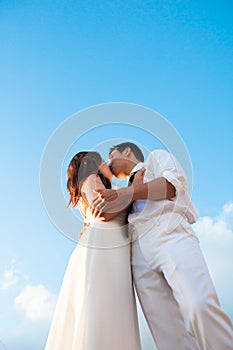 Romantic couple dressed in white, kissing under the blue sky on their wedding day.