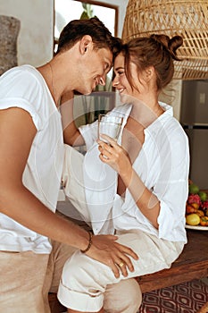 Romantic Couple Drinking Water At Kitchen. Beautiful Woman And Handsome Man Sitting On Wooden Table And Looking Lovely.