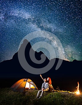 Romantic couple hikers looking at the shines starry sky at night. Happy pair sitting near camp and campfire