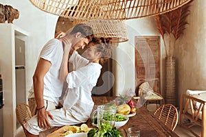 Romantic Couple At Kitchen. Beautiful Woman Sitting On Wooden Table And Hugging Standing Man.