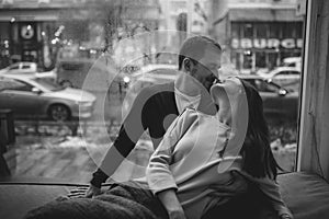 Romantic couple. Loving guy kisses his beautiful girlfriend sitting on the windowsill in a cozy cafe. Black and white