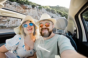 Romantic couple making selfie on smartphone camera in rental cabrio car on ocean or sea beach enjoying summer vacation