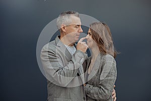 Romantic couple, mature gray haired man and young woman hugging, smiling and touching nose against gray wall outdoors