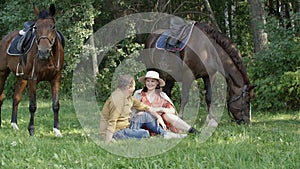 Romantic couple sitting on green grass on grazing horses background. Happy man and woman relaxing on rural pasture