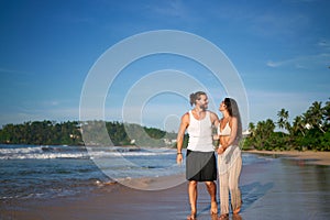 Romantic couple strolls on tropical beach at sunset. Man and woman in love walk barefoot by sea, enjoy honeymoon