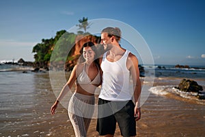 Romantic couple strolls on tropical beach at sunset. Newlyweds on honeymoon enjoy seaside walk. Man, woman in casual