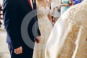 Romantic couple, stylish bride and handsome groom holding hands in church during wedding ceremony in catholic chapel close-up,