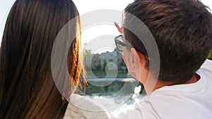 Romantic couple talking and sitting on the green grass near the pond on sunset. Back view. Couple trust, love and