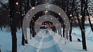 romantic couple walking by illuminated alley in snowy winter park, snow in forest and road with decorated light bulbs 4k