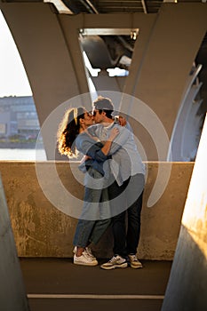 Romantic couple of young male and female embracing and kissing at sunset standing under urban bridge