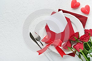 Romantic creative table setting. Empty white plate and red roses, candles, knife, fork and decorative silk hearts on white