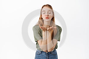 Romantic cute girlfriend sending air kiss at camera with closed eyes, puckered lips and hands near mouth, standing over