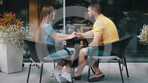 Romantic date of young happy couple in love smiling talking sitting at table in cafe outside on summer day, medium view