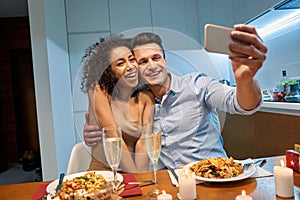 Romantic Date. Young multiethnic couple at home having dinner hugging taking selfie on smartphone saving moment laughing