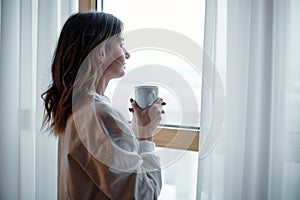 Romantic dreamy female homeowner tenant holding mug of hot coffee looking out of window. Leisurely morning