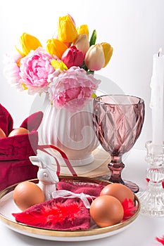 Romantic Easter table setting with candles, white plate, pink wine glass Table decoration with rabbit and painted eggs in red and