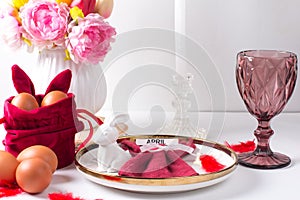 Romantic Easter table setting with candles, white plate, pink wine glass Table decoration with rabbit and painted eggs in red and